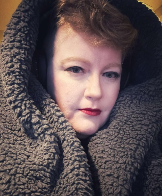 A redhead femme with blue eyes wrapped up in a warm fleece hood.