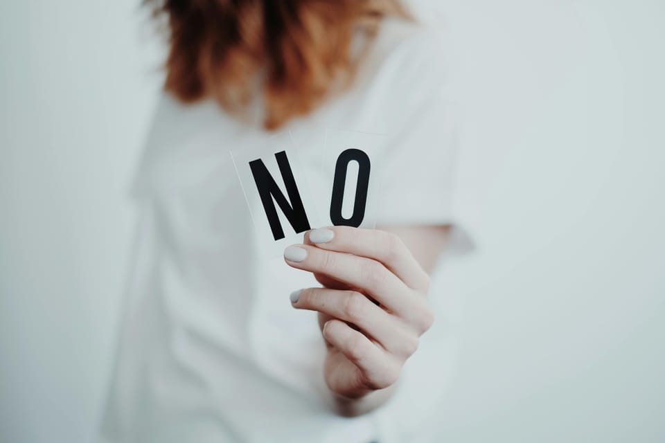 A person wearing white, standing in front of a white background. In focus, their hand holds up the word "No."
